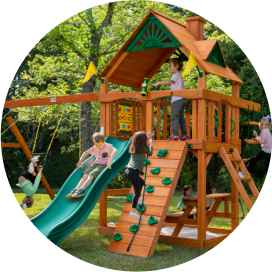 Kids playing on a wooden playset with a slide, swings, a climbing wall, rope ladder and clubhouse.