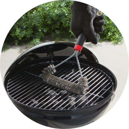 A gloved hand running a grill brush across the grates of a small black charcoal grill on a patio.