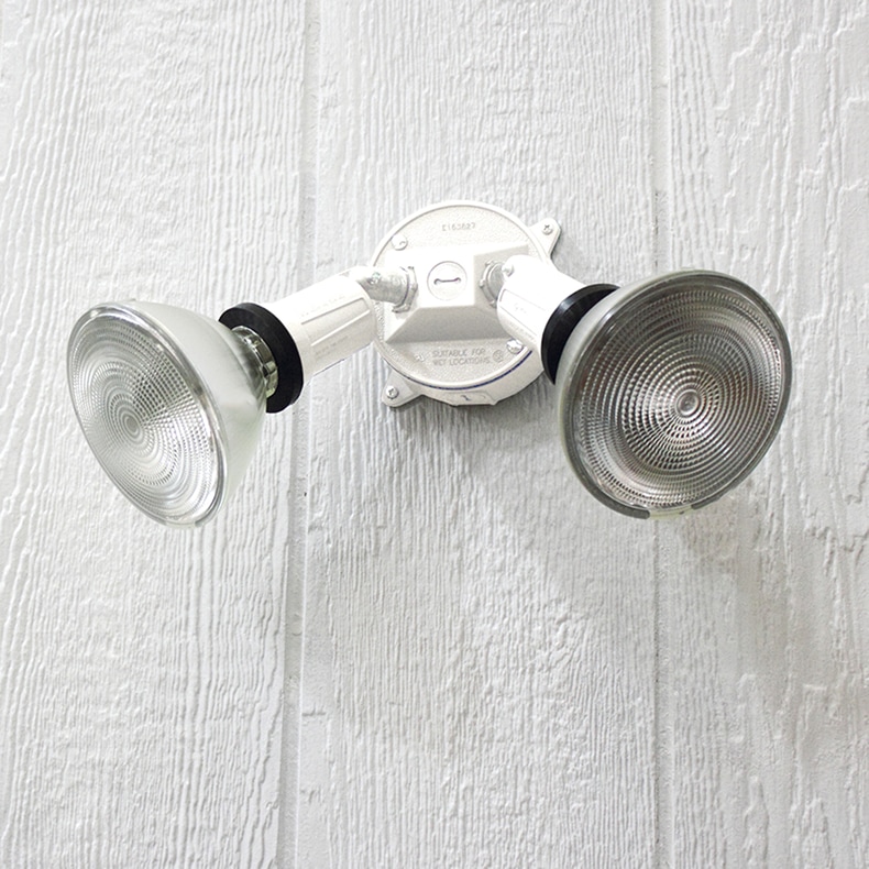 A white switch-controlled two-head incandescent flood light mounted on the side of a house at night.