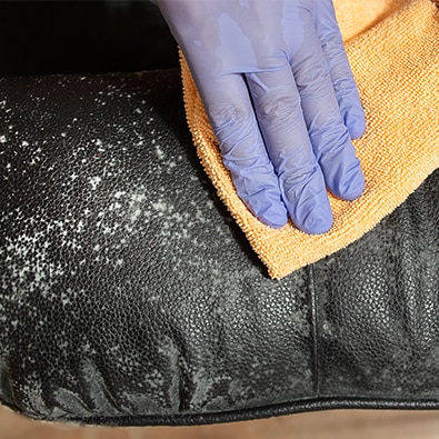 A person wearing cleaning gloves wiping mildew off of a black leather chair with a yellow towel.