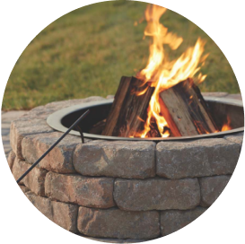 A fire pit made from a kit with beige concrete pavers that has a log burning inside.