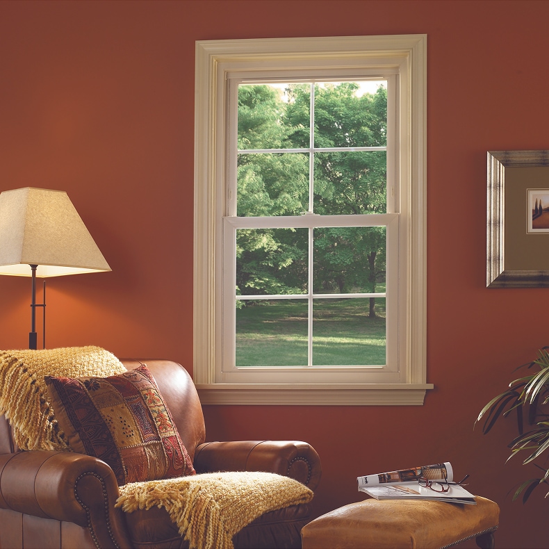 A room with a dark patterned rug, brown leather chair, wood table, guitar and white windows.
