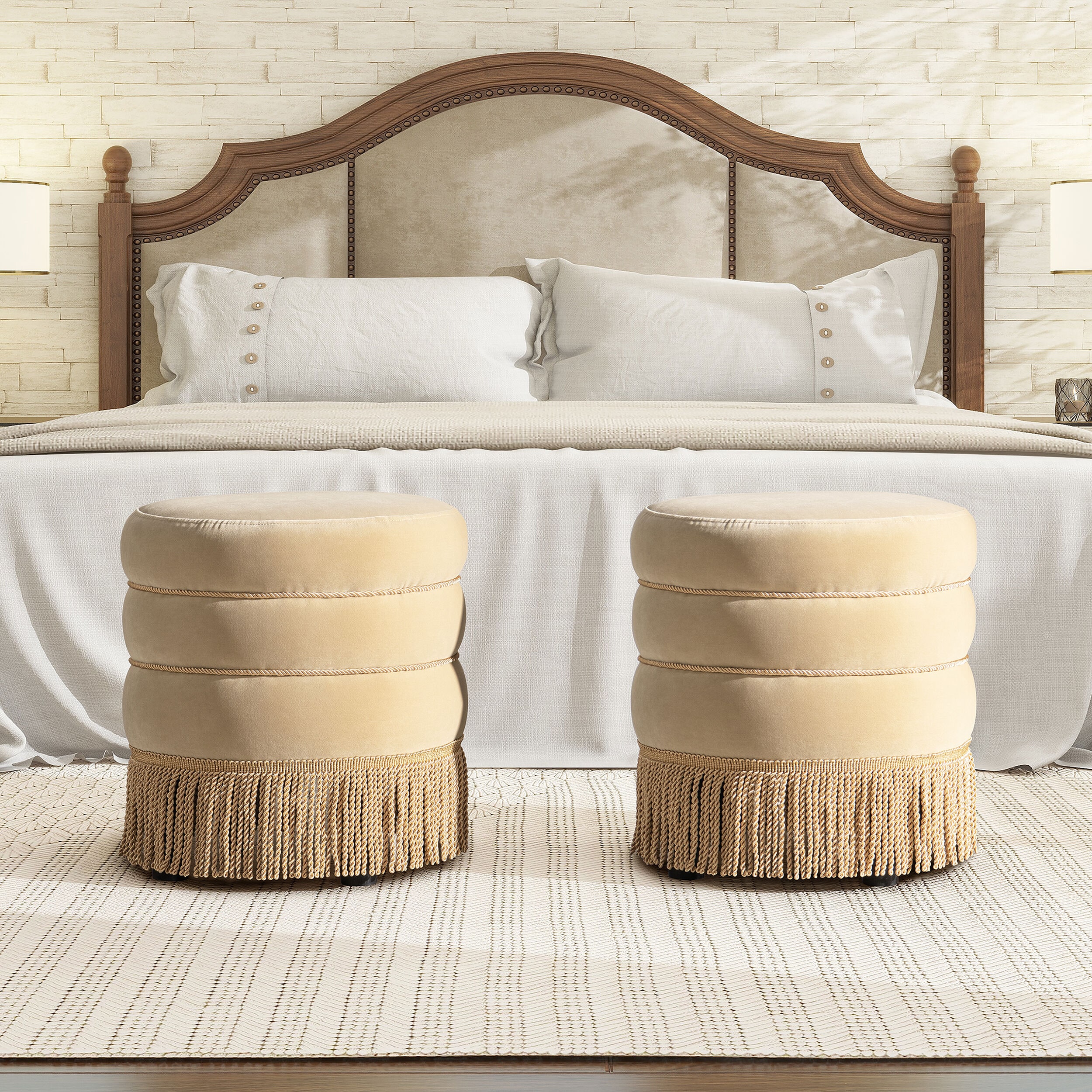 Two beige fringe-trim round ottomans in front of a bed in a bedroom scene